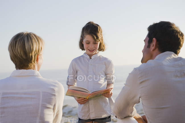 Girl reading book in front of parents on beach — Stock Photo