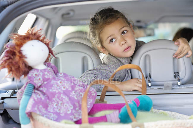 Menina bonito sentado no carro com boneca e olhando para a janela traseira — Fotografia de Stock