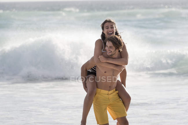 Glückliche junge Frau reitet huckepack auf den Schultern ihres Freundes am Strand — Stockfoto
