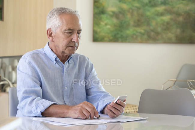 Senior benutzt Handy bei Papierkram zu Hause — Stockfoto