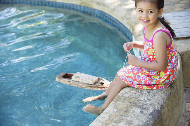 Girl sitting at edge of swimming pool with toy boat in water — Stock Photo