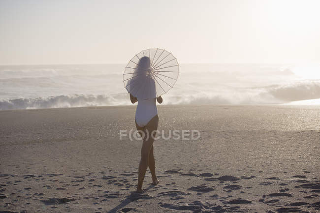 Femme mince en maillot de bain marchant sur la plage avec parasol — Photo de stock