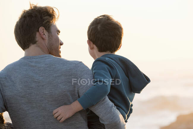 Feliz padre e hijo de pie en la playa al atardecer - foto de stock