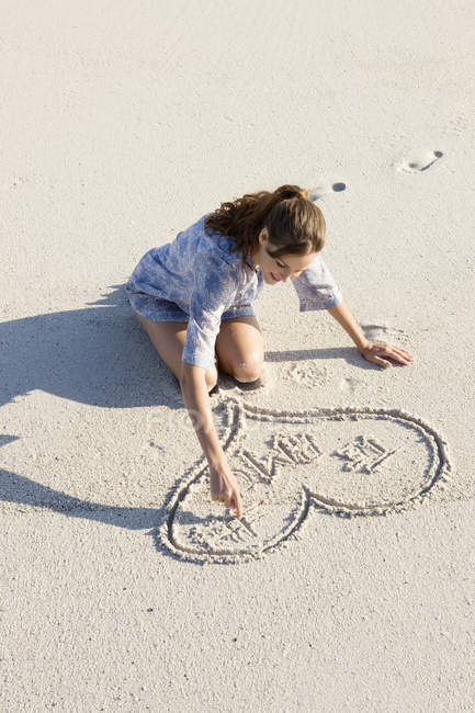 Smiling young woman drawing heart shape on sandy beach — Stock Photo