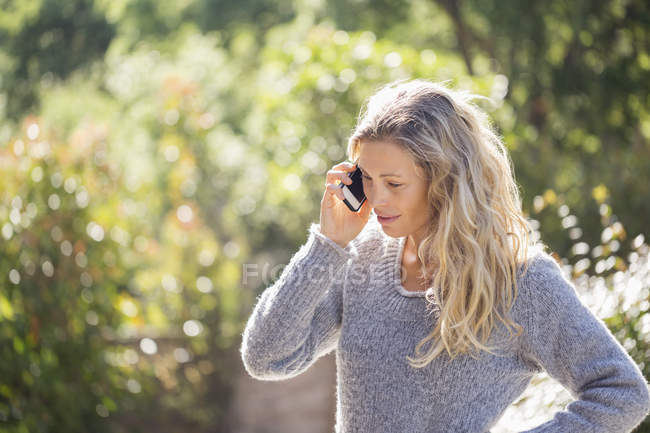 Woman in sweater talking on phone in sunny garden — Stock Photo