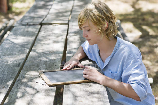 Teenage boy writing on slate at wooden table outdoors — Stock Photo