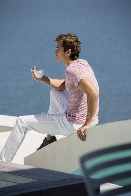Jeune homme dégustant une tasse de café sur la terrasse avec vue sur la mer — Photo de stock