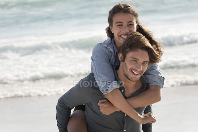 Feliz joven mujer cabalgando a cuestas en los hombros novio en la playa - foto de stock