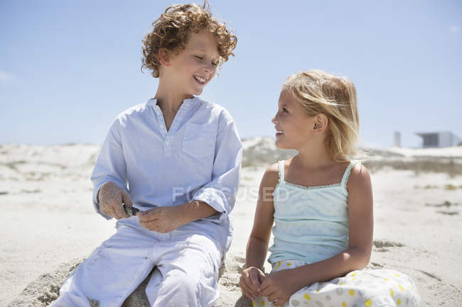 Niño con hermana sentada en la playa de arena y sonriendo - foto de stock
