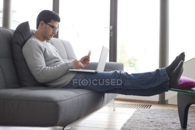 Homem segurando cartão de crédito e usando um laptop no sofá — Fotografia de Stock