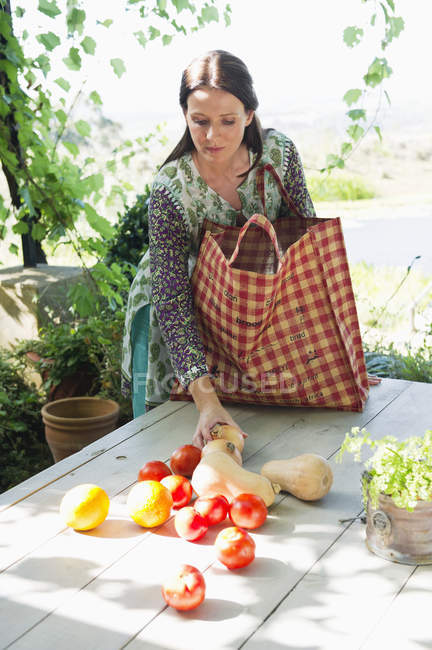 Mature woman collecting vegetables and fruits from wooden table in garden — Stock Photo