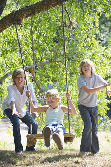 Усміхнені брати і сестри грають в гойдалки на дереві в літньому саду — стокове фото