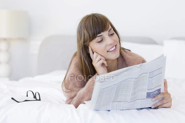Улыбающаяся молодая женщина читает газету на кровати — стоковое фото
