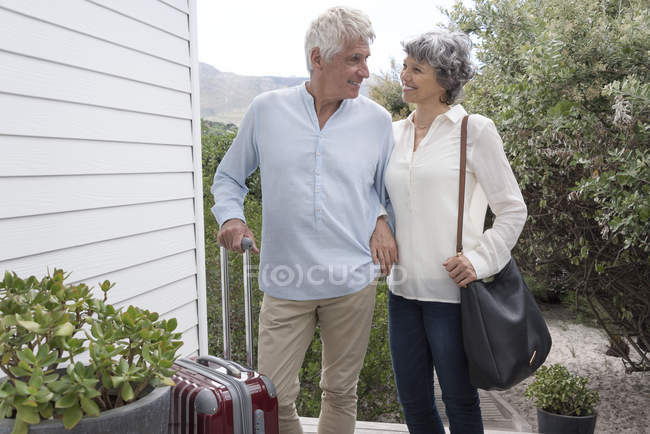 Happy senior couple standing with suitcase outside of house — Stock Photo