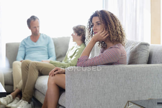 Mujer aburrida sentada en el sofá en la sala de estar con el marido y el hijo en el fondo - foto de stock
