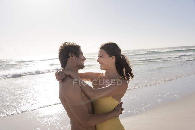 Sorrindo jovem casal abraçando na praia ensolarada — Fotografia de Stock