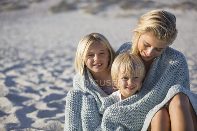 Портрет улыбающейся матери и детей, завернутых в шаль, сидящих на пляже — стоковое фото
