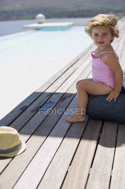 Happy baby girl sitting on boardwalk near infinity pool and looking at camera — Stock Photo