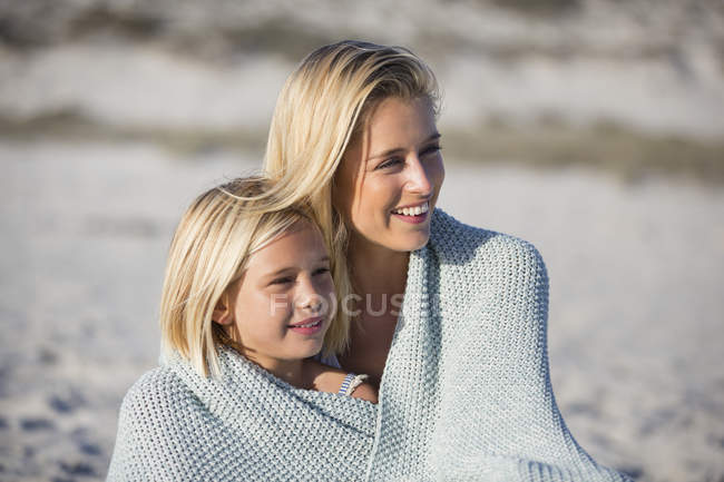 Primer plano de la sonriente rubia y su hija sentadas en la playa de arena envueltas en una toalla - foto de stock