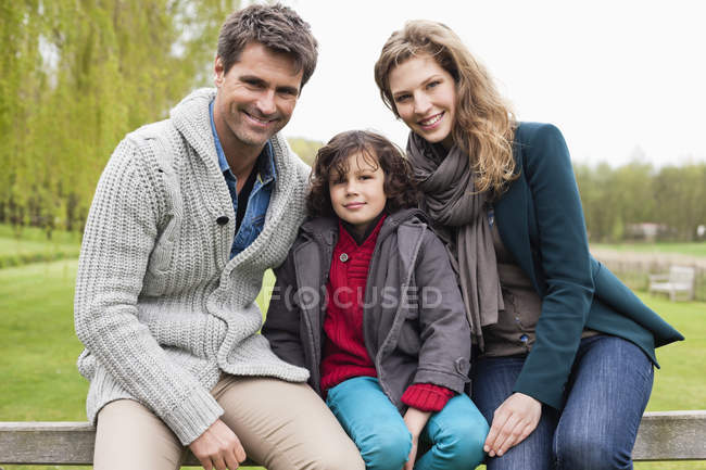 Portrait of boy sitting with parents on wooden fence in countryside — Stock Photo