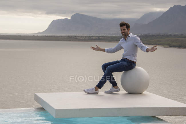 Man sitting on stone ball and gesturing at lake shore — Stock Photo