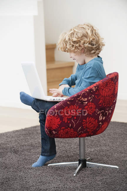 Lindo niño sonriente con el pelo rubio usando un ordenador portátil en sillón en casa - foto de stock