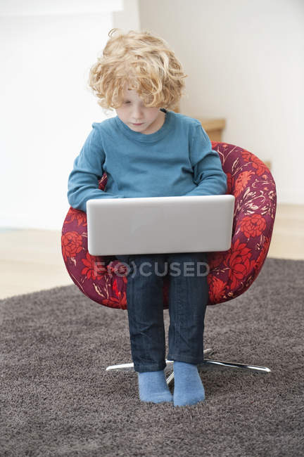 Lindo chico con pelo rubio usando un portátil en sillón en casa - foto de stock