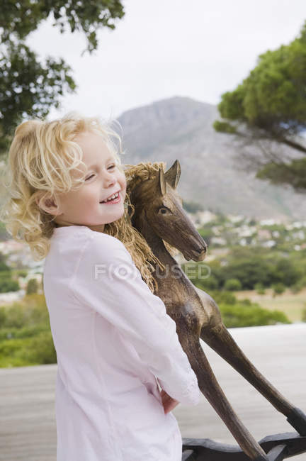 Niña sonriente jugando con un caballo mecedora en la naturaleza - foto de stock