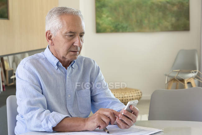 Старший чоловік використовує мобільний телефон, роблячи документи вдома — стокове фото
