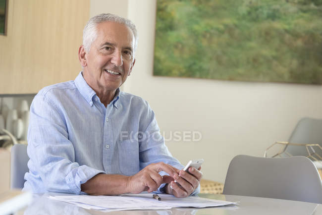 Retrato de homem idoso feliz usando telefone celular enquanto fazendo papelada em casa — Fotografia de Stock