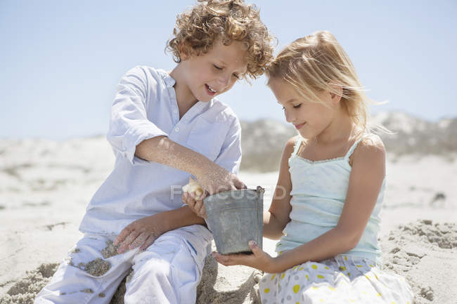 Niño con hermana sentado en la playa de arena y jugando - foto de stock