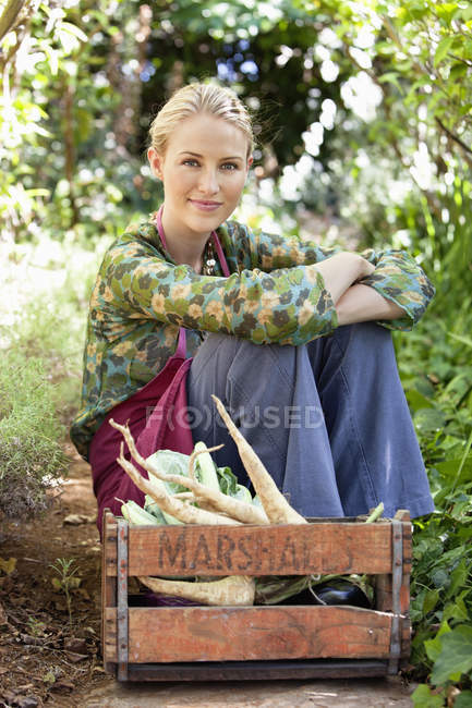 Улыбающаяся женщина сидит рядом с ящиком свежих собранных овощей в саду — стоковое фото