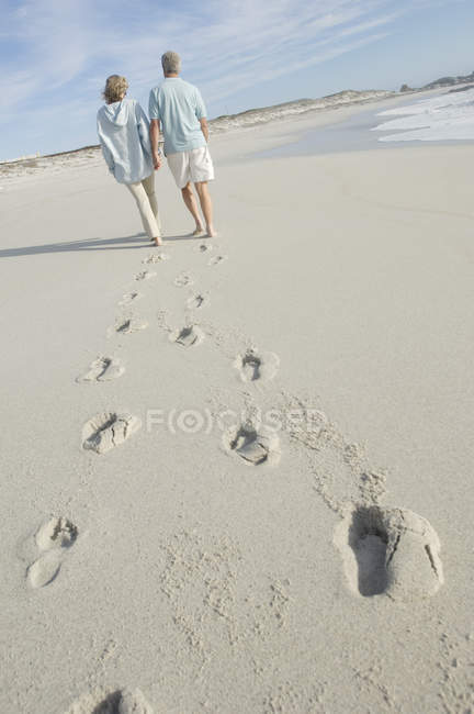 Vista trasera de pareja adulta caminando en la playa cogidos de la mano - foto de stock