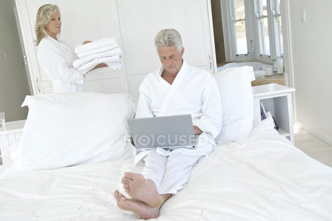 Pareja en el dormitorio, mujer llevando pila de toallas, hombre usando el ordenador portátil - foto de stock