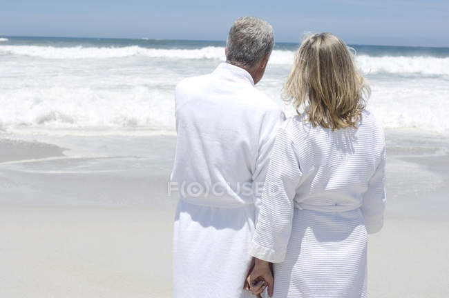 Vista trasera de la pareja en albornoces de pie en la playa y mirando a la vista - foto de stock