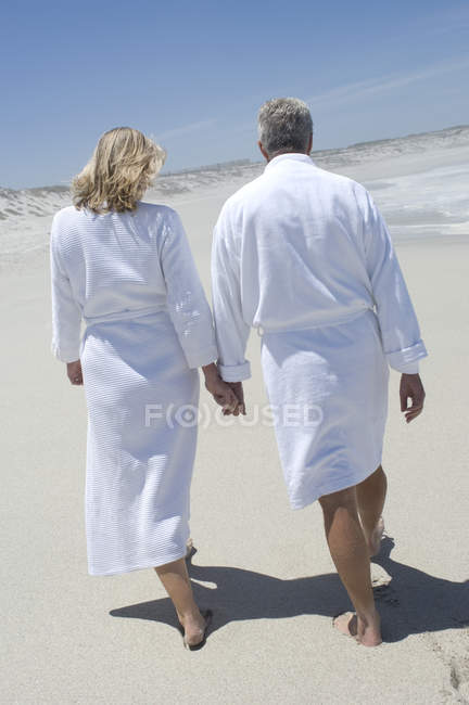 Vista posteriore di coppia in accappatoi passeggiando sulla spiaggia sabbiosa tenendosi per mano — Foto stock