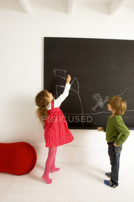 Menina desenhando em um quadro negro e seu irmão observando-a — Fotografia de Stock