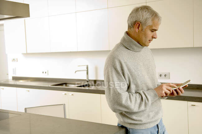 Hombre maduro de pie en la cocina y mensajes de texto en un teléfono móvil - foto de stock
