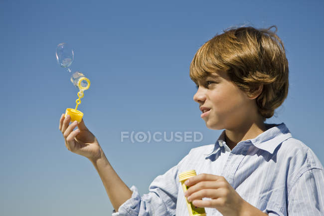 Niño sosteniendo una varita de burbuja contra el cielo azul - foto de stock