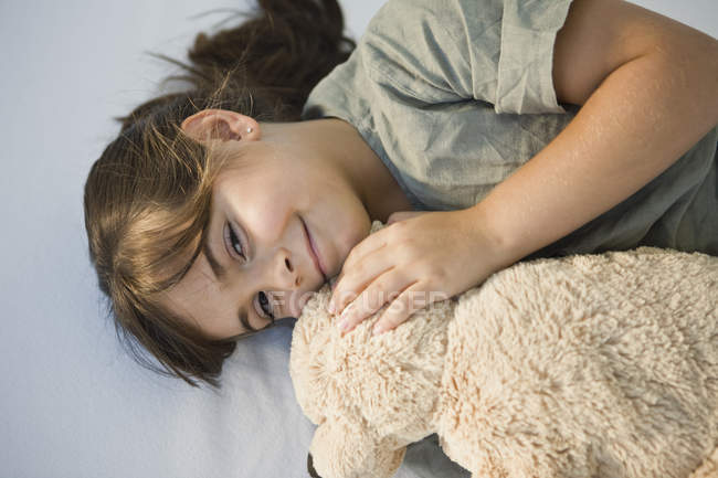 Retrato de una linda niña sonriente acostada con un oso de peluche en la cama - foto de stock