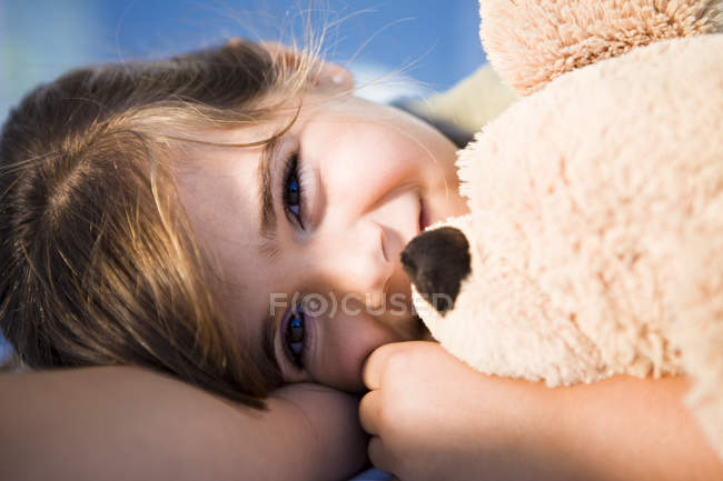 Усміхнена маленька дівчинка, що качає плюшевого ведмедя на сонячному світлі — стокове фото