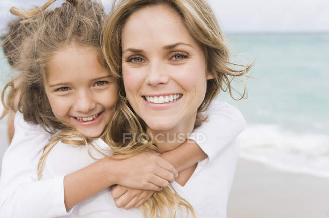 Mulher dando filha piggyback na praia e olhando para a câmera — Fotografia de Stock