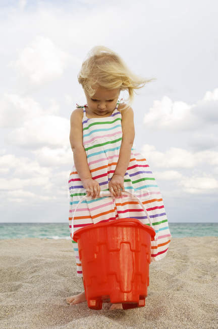Menina bonito segurando balde de areia na praia e olhando para baixo — Fotografia de Stock