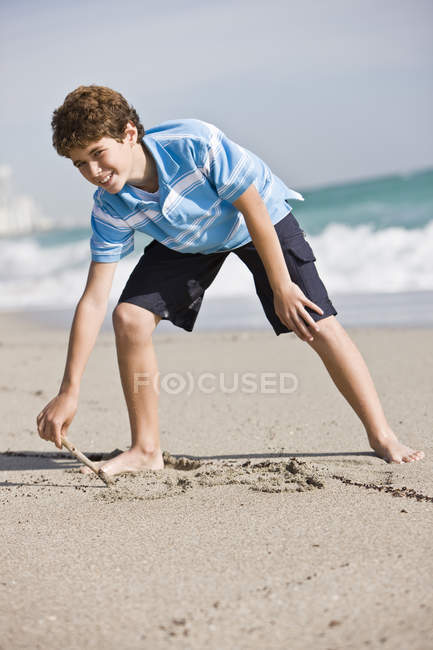 Мальчик-подросток рисует в песке на солнечном пляже — стоковое фото