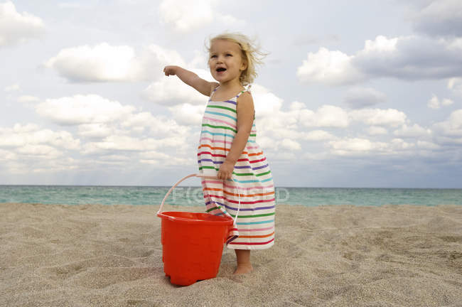 Милая маленькая девочка держит ведро с песком на пляже и указывает на море — стоковое фото