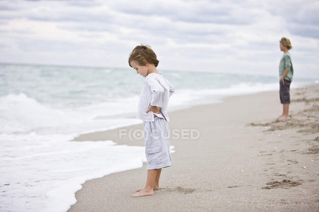 Маленький хлопчик стоїть на піщаному пляжі з братом на фоні — стокове фото