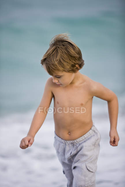 Menino sem camisa andando na praia e olhando para baixo — Fotografia de Stock
