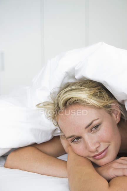Nahaufnahme einer verträumten Frau, die unter einer Bettdecke auf dem Bett liegt und lächelt — Stockfoto