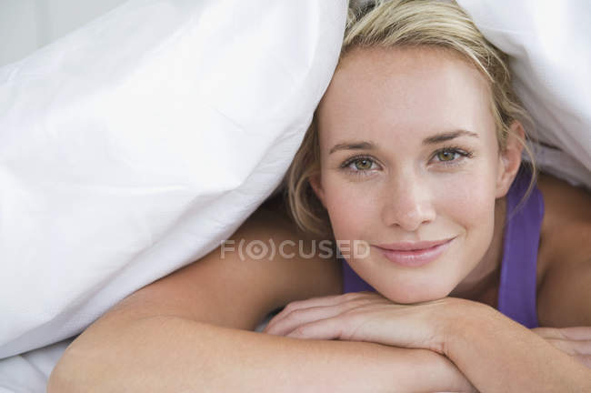 Primo piano della donna sdraiata sul letto sotto il piumone e sorridente — Foto stock