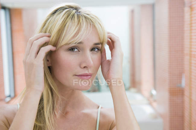 Blonde jeune femme ajuster les cheveux devant le miroir dans la salle de bain — Photo de stock